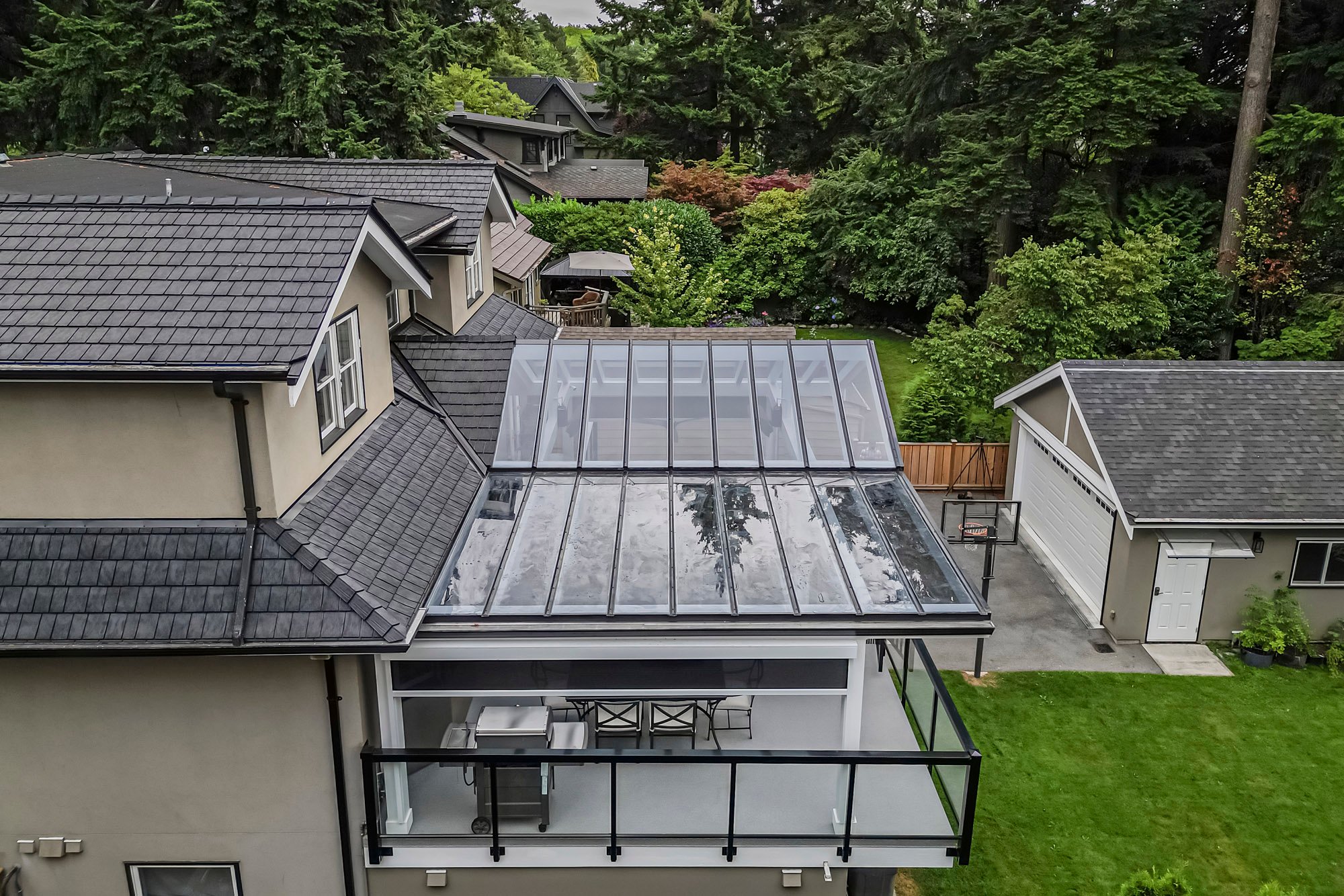 Looking down at the glass-paneled roof of a deck attached to a house, with a grill and dining table visible underneath.