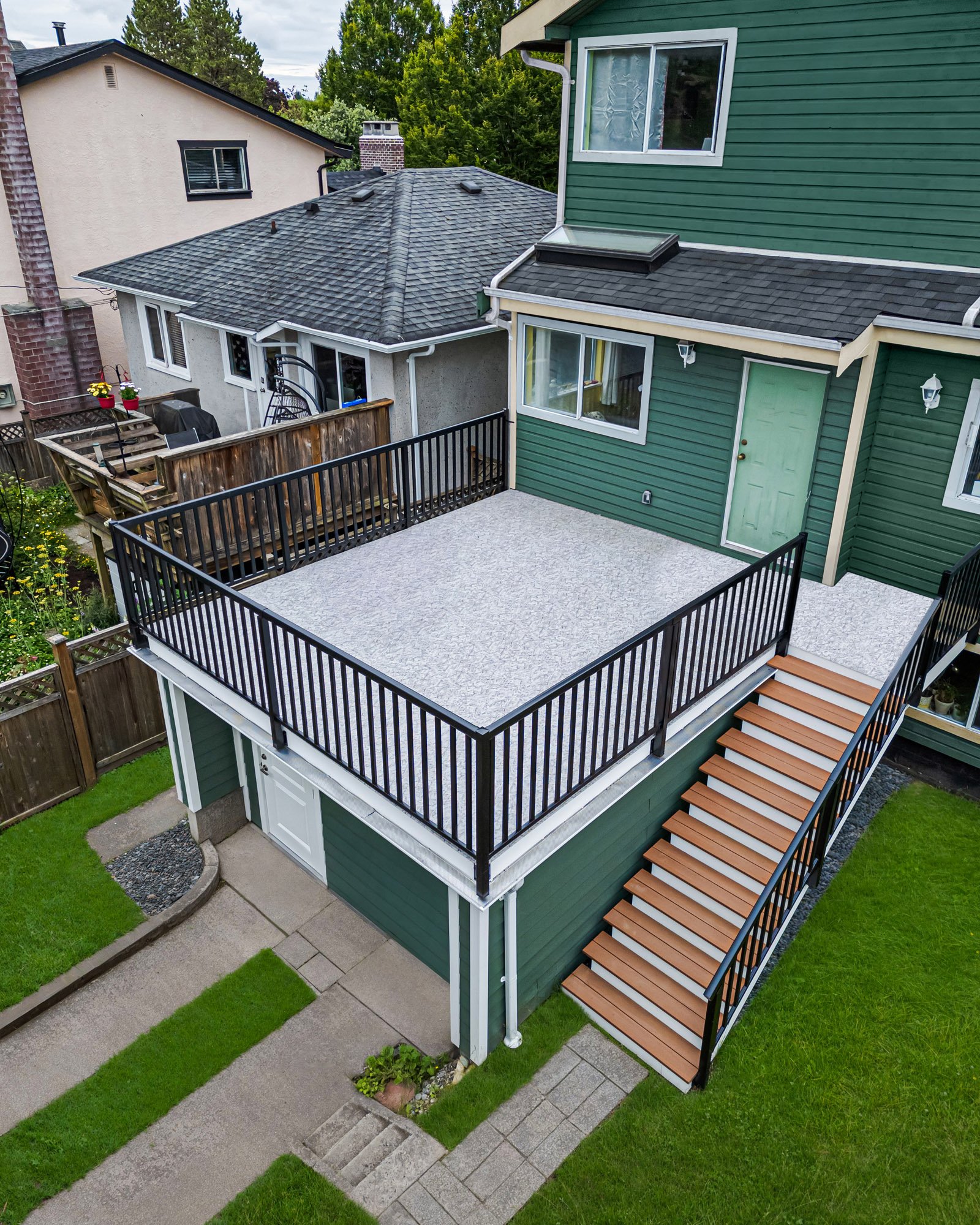 High-angle view of a green residential home with a flat roof deck, black railings, and wood stairs leading to the backyard.