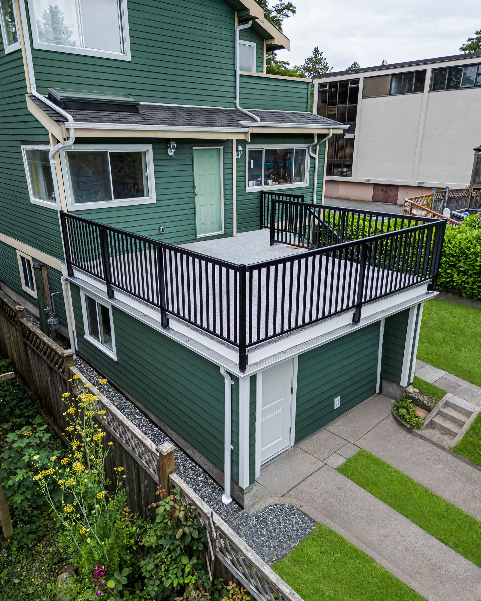 Aerial view of a green house exterior featuring a flat roof deck with black metal railings above an enclosed lower level.