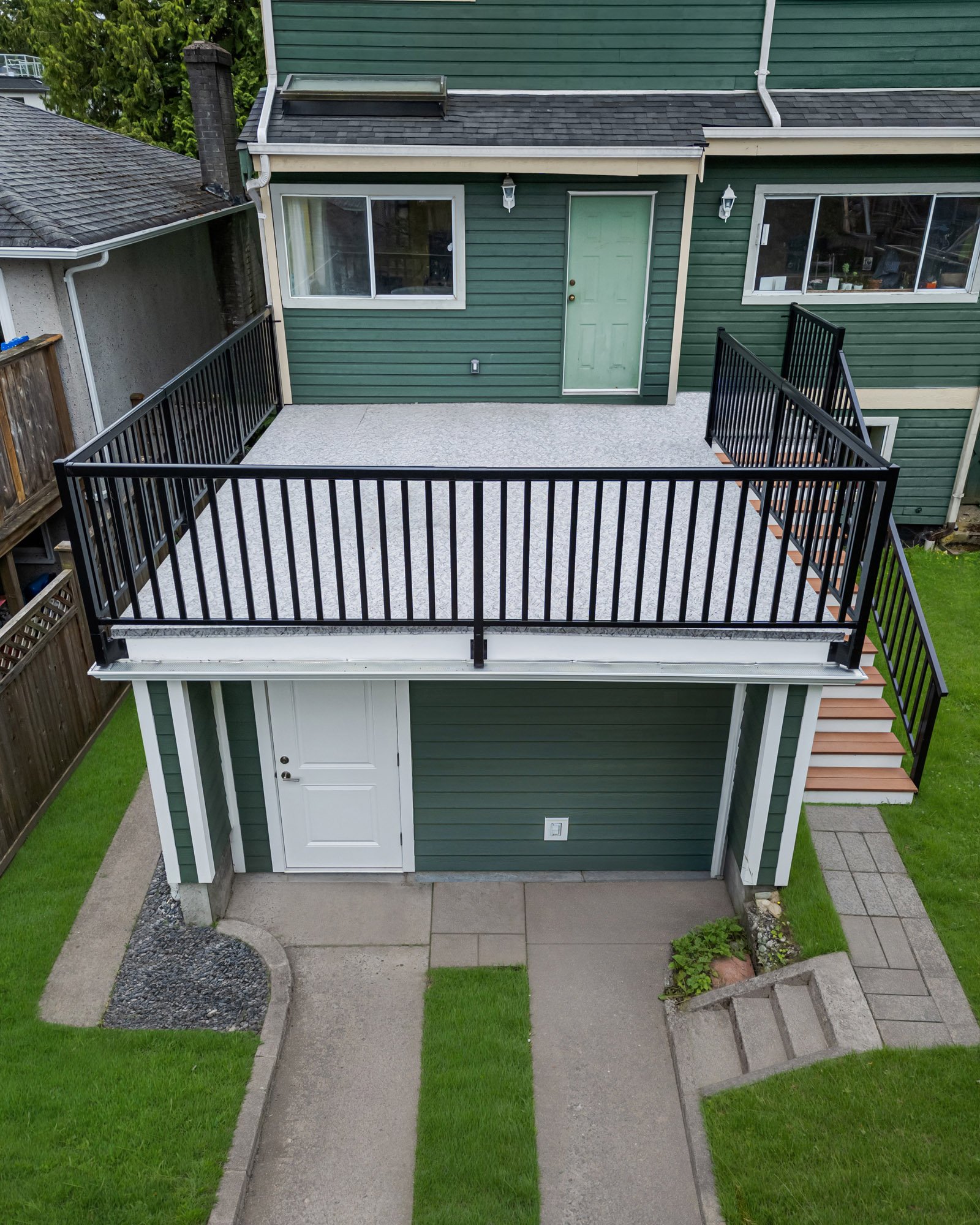 Straight-down aerial shot of a grey flat roof deck with black metal railings built over a lower green siding house exterior.