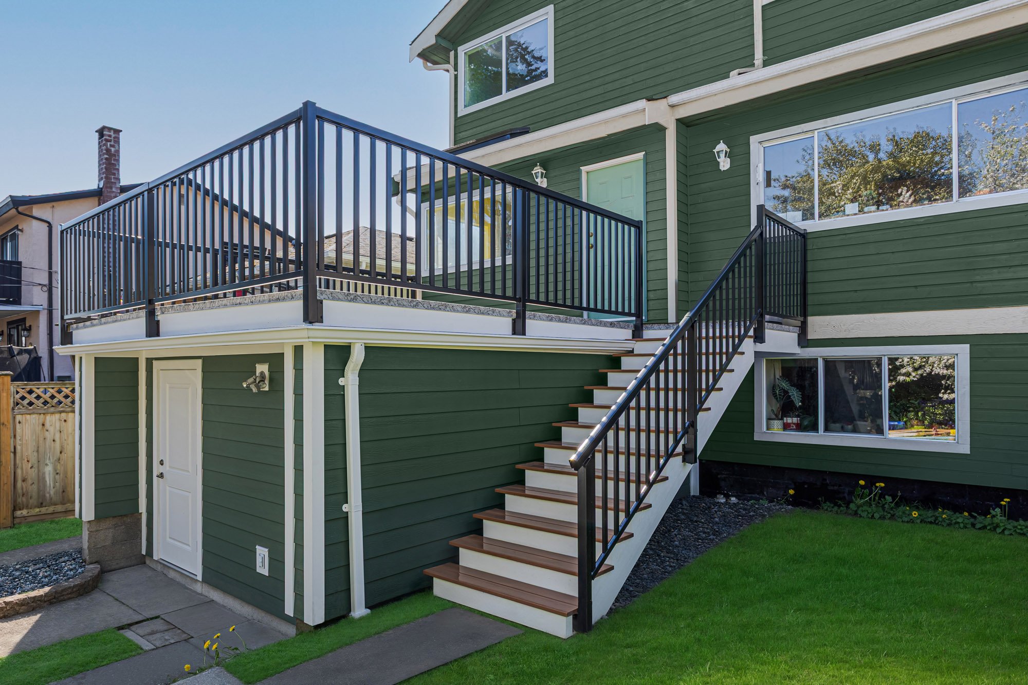 Exterior view of a green house featuring a flat roof deck with black railings and wooden steps leading down to a green lawn.