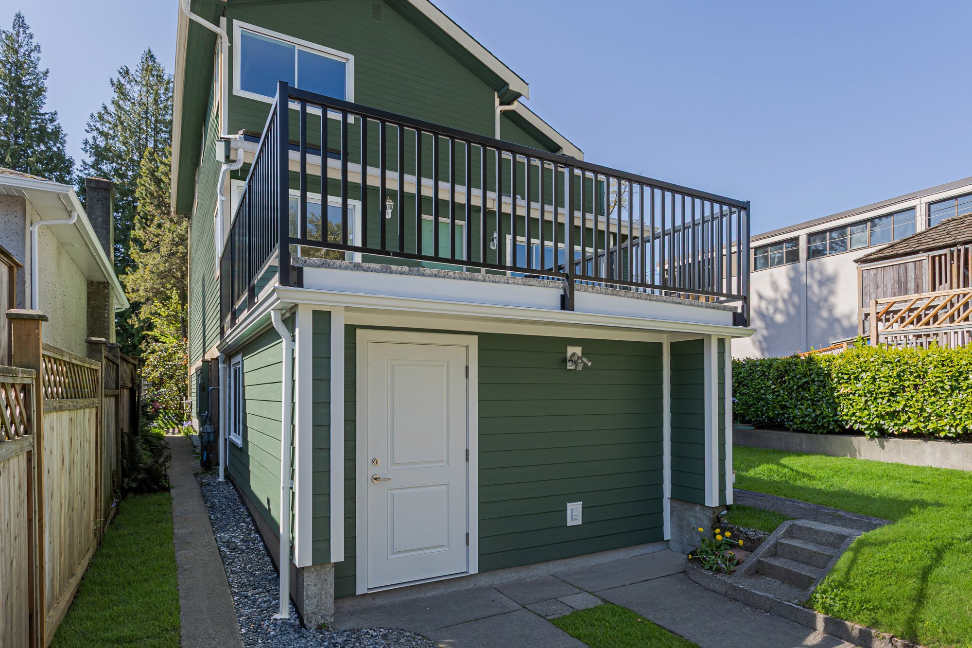 Side view of a green siding house displaying a flat roof deck with black railings situated above a ground-level white door.