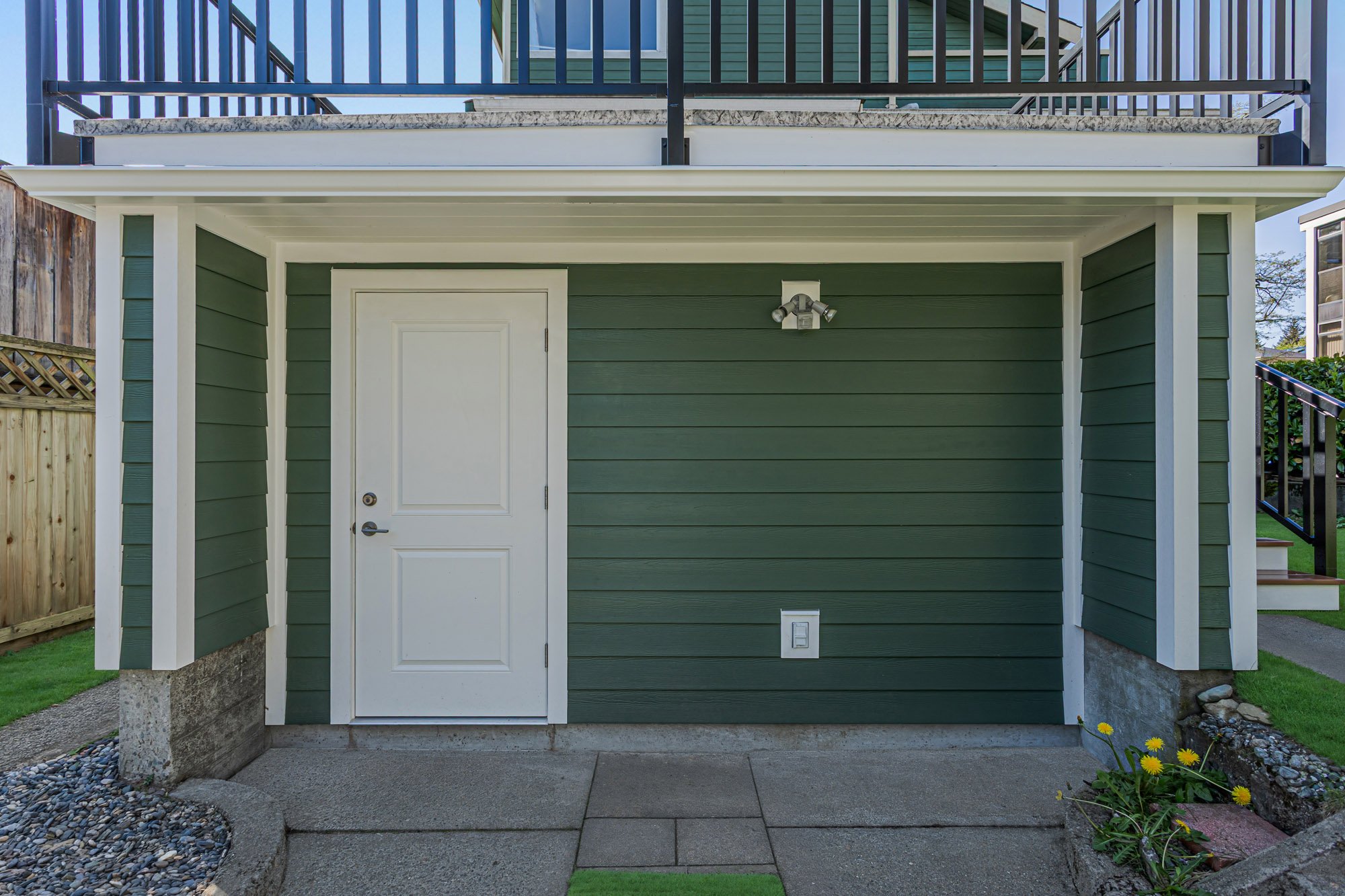 Close-up of the ground level exterior of a green house, featuring a white entry door with silver hardware below a raised deck.