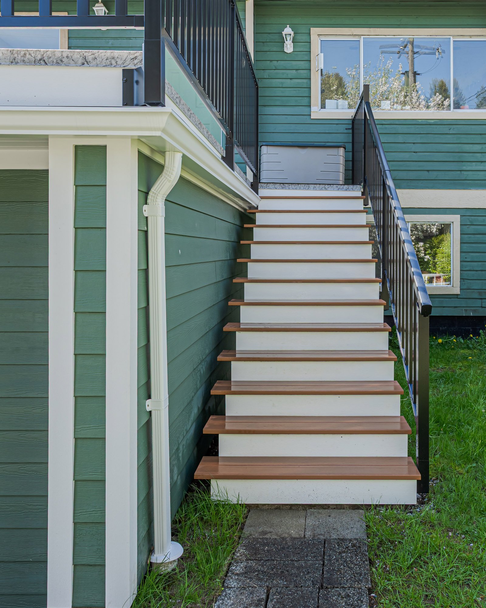Looking straight up a set of outdoor stairs with brown wood treads and white risers beside a green house with black railings.