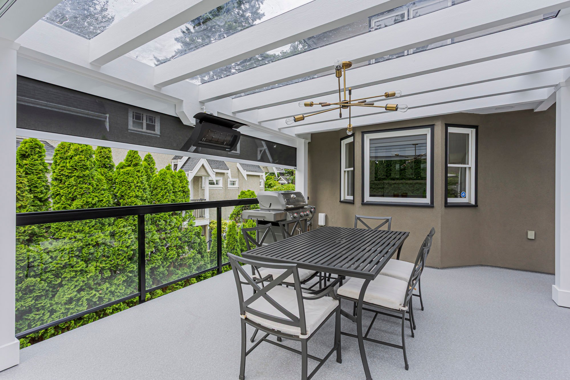 A covered outdoor deck featuring a glass roof, a grey dining table with six chairs, and a built-in grill area.