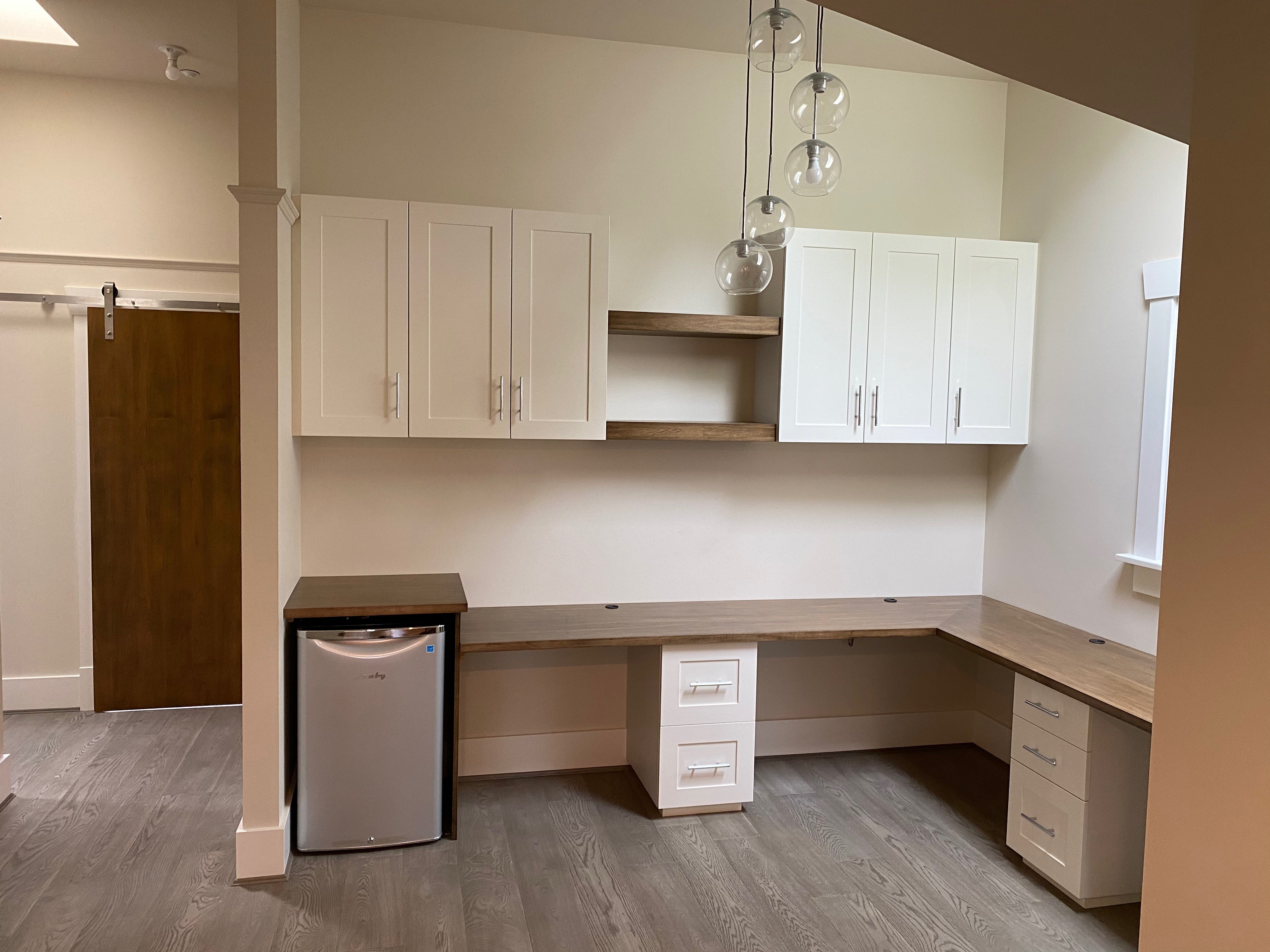 A home office area with a built-in L-shaped wood desk, white upper cabinets, floating wood shelves, and a small silver fridge.