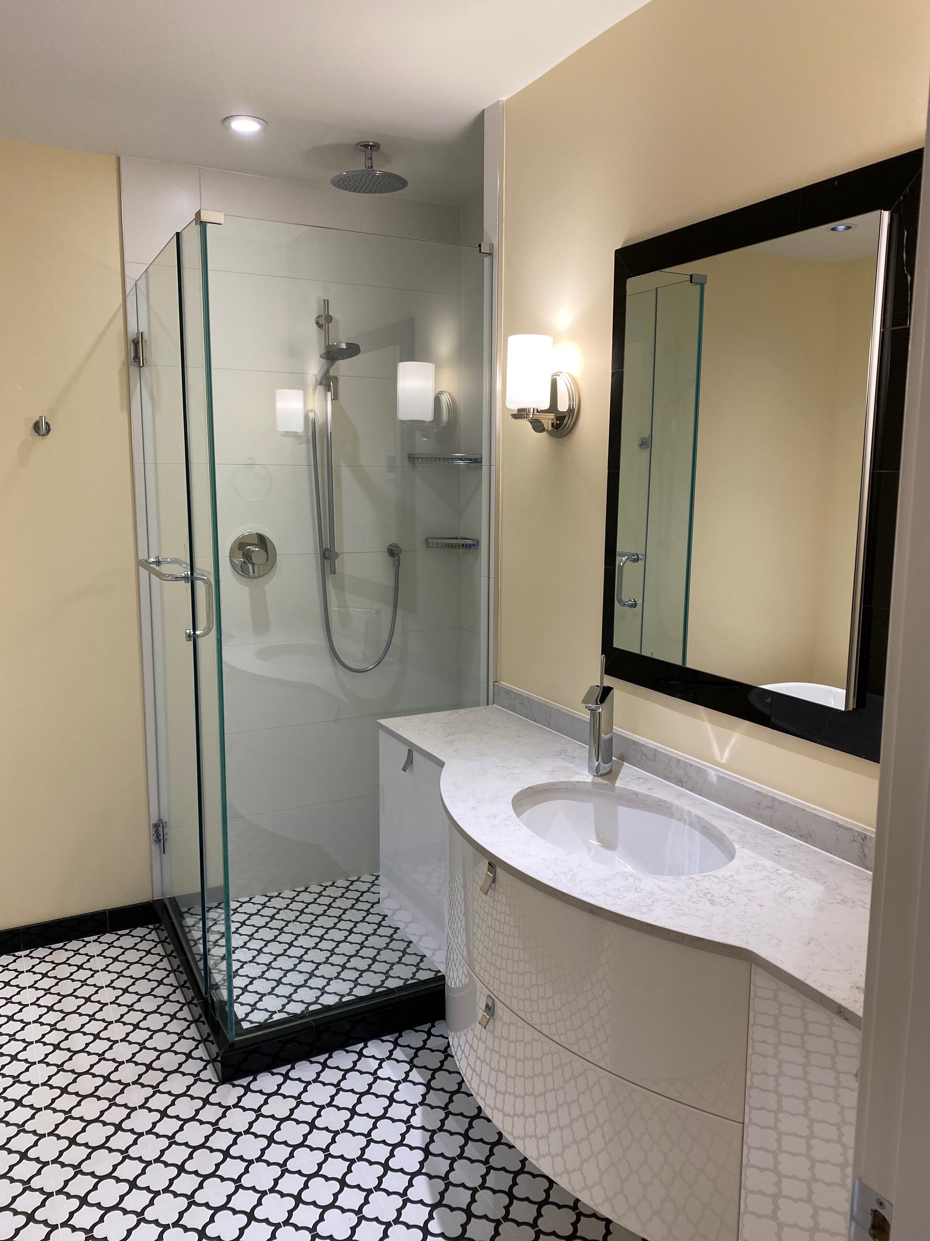 A bright bathroom with a curved white vanity cabinet, glass shower stall, a toilet, and black and white patterned tile.