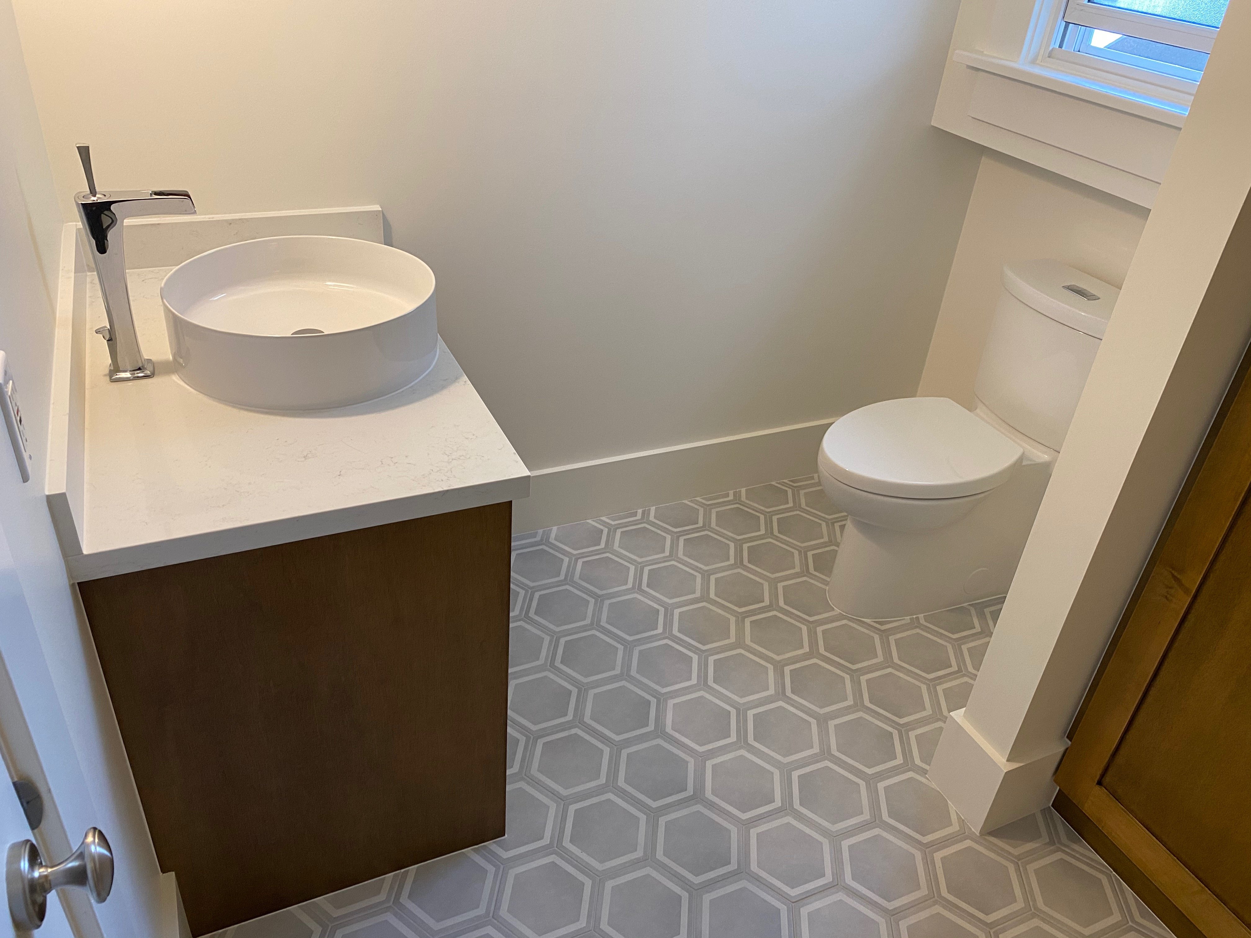 A small modern bathroom featuring a wood vanity with a white vessel sink, a standard toilet, and grey hexagonal floor tiles.