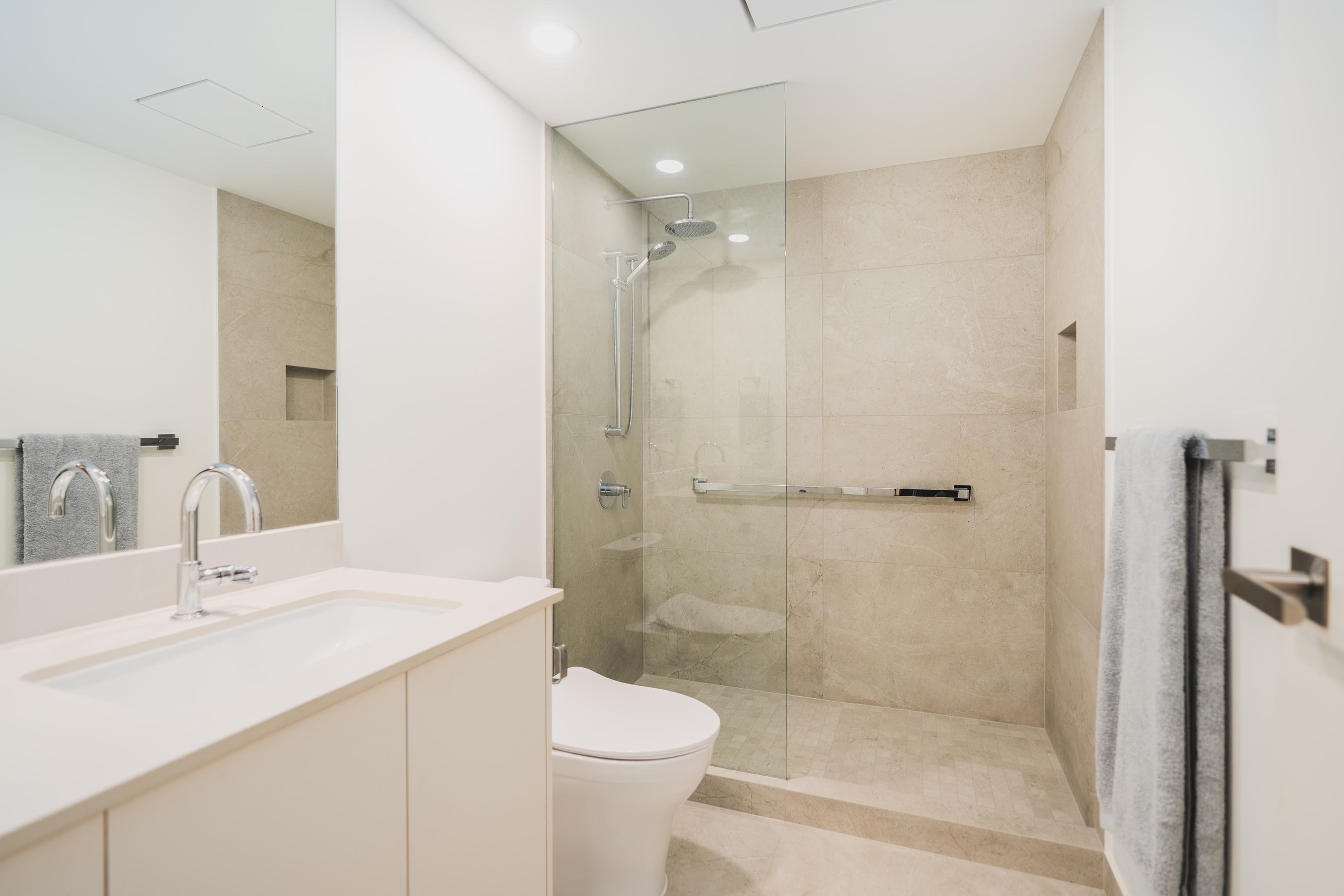 A bright modern bathroom featuring a white vanity with an under-mount sink, a white toilet, and a beige tiled glass shower.