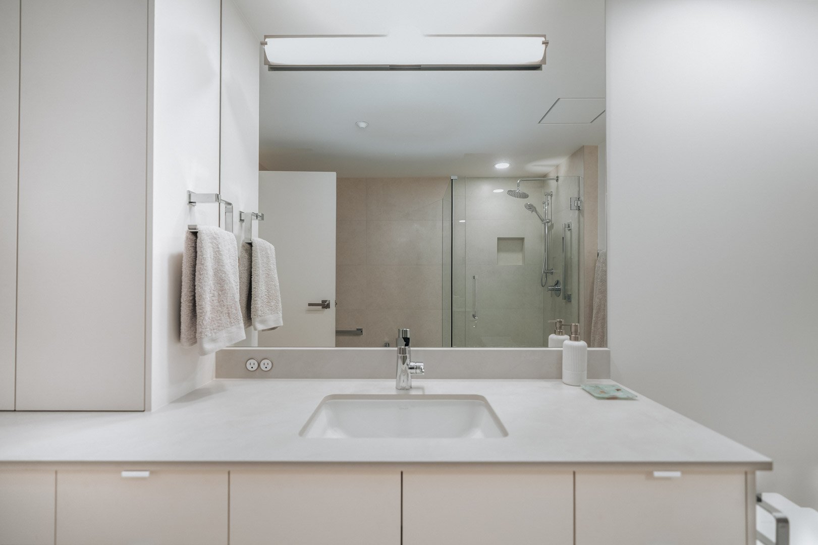 A modern bathroom with white vanity cabinets, a large mirror, a white toilet, and a doorway leading to an adjacent carpeted room.
