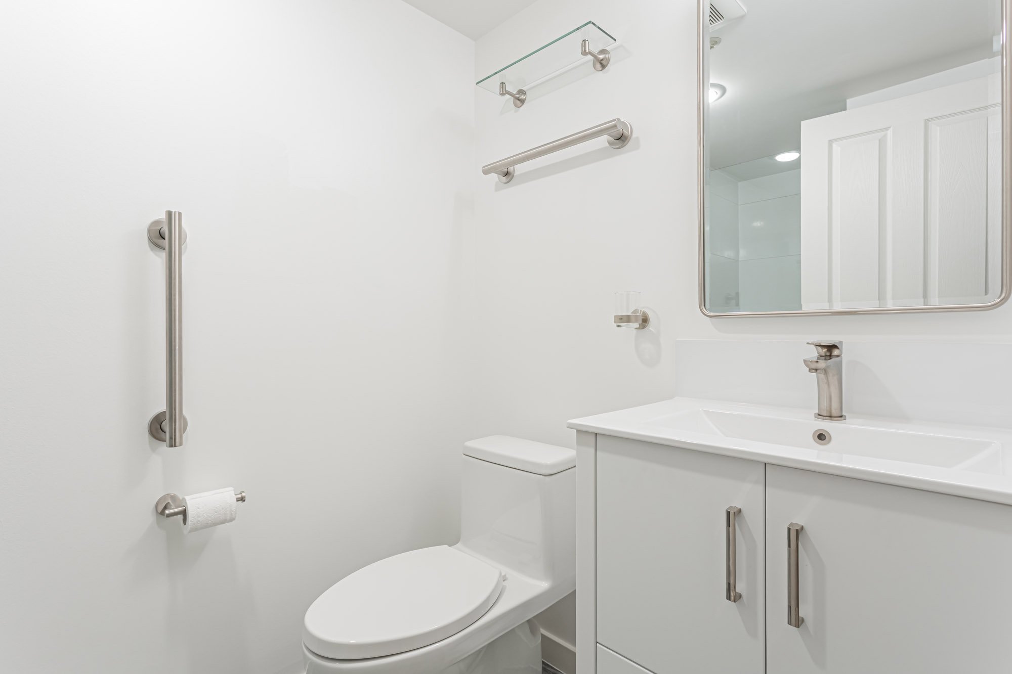 A bright white accessible bathroom featuring a toilet, wall-mounted grab bars, a glass shelf, and a modern vanity cabinet.