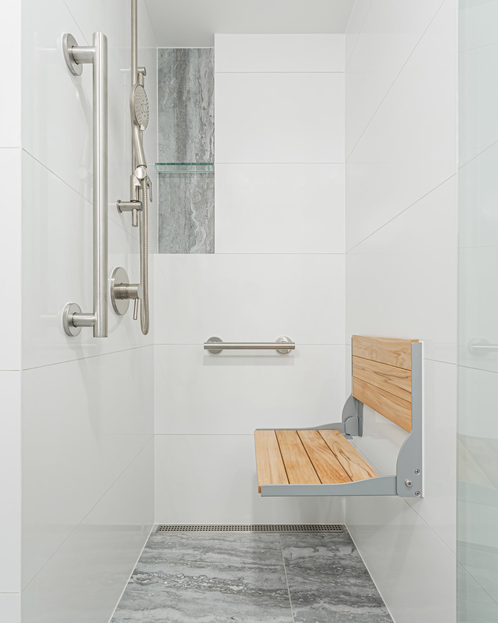 A bright white shower stall featuring a grey marble accent niche, brushed nickel grab bars, and a folding wooden bench seat.