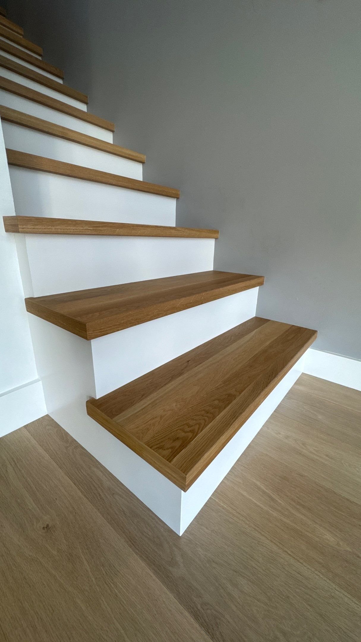 Side angle view looking up a modern staircase with light oak wood treads and solid white painted risers beside a grey wall.