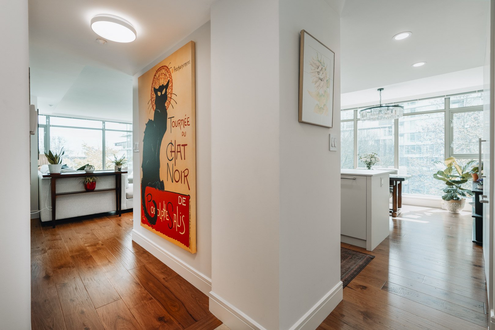 A bright hallway with warm hardwood floors featuring a large poster of a black cat, leading toward a sunlit living area.