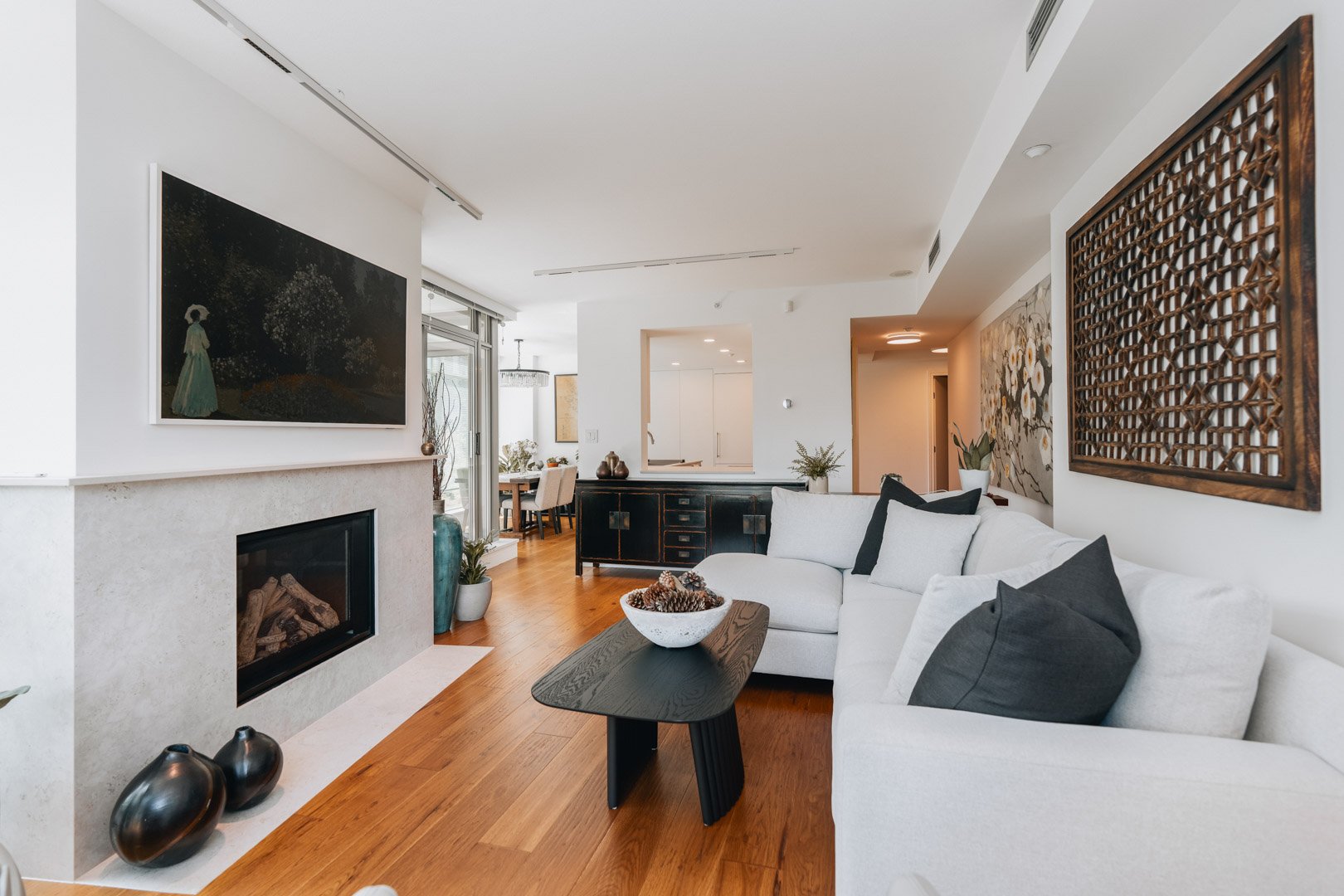 A bright modern living room featuring a white sectional sofa, black coffee table, stone fireplace, and warm hardwood floors.
