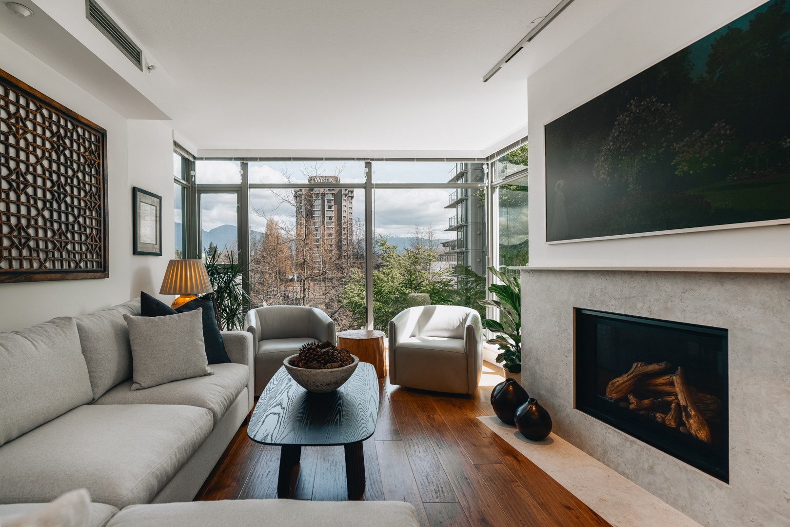 A spacious, bright living room featuring a light stone fireplace, a large grey sectional sofa, and expansive corner windows.