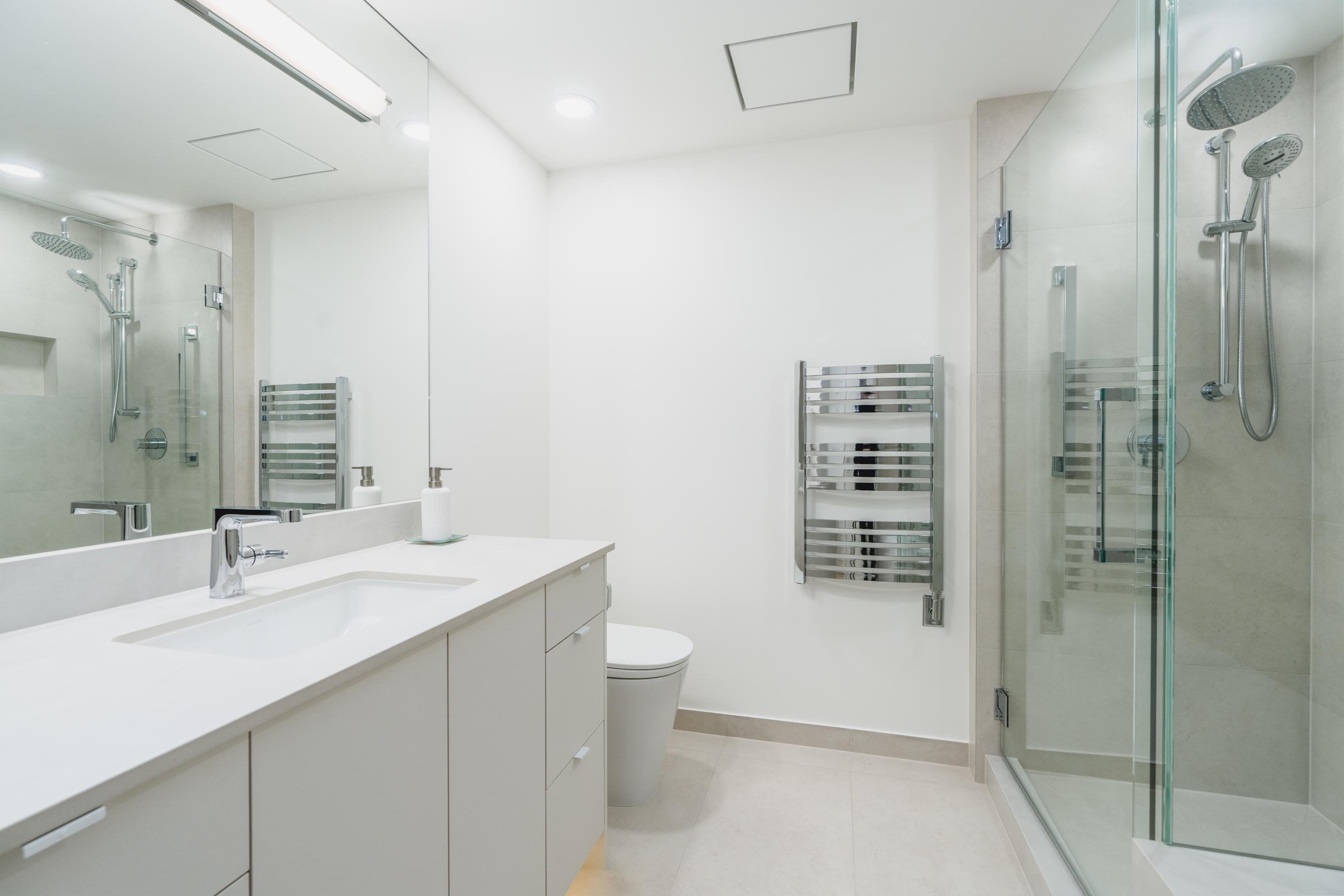 View inside a bright bathroom with a white vanity sink, a large mirror reflecting the shower stall, and a chrome towel warmer.