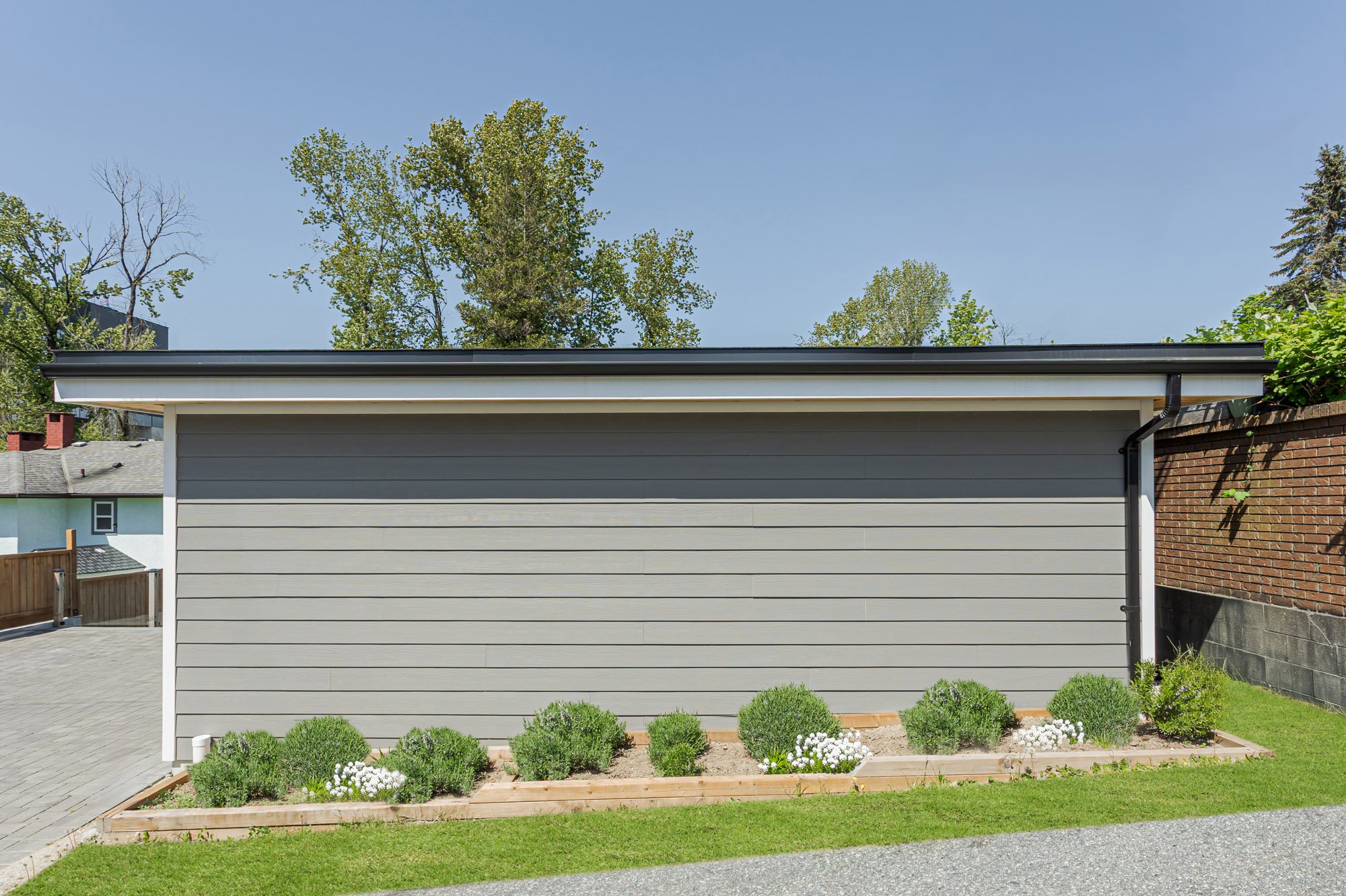 Side exterior view of a detached garage featuring long grey wood siding, a flat roof, and a low planter box with green shrubs.