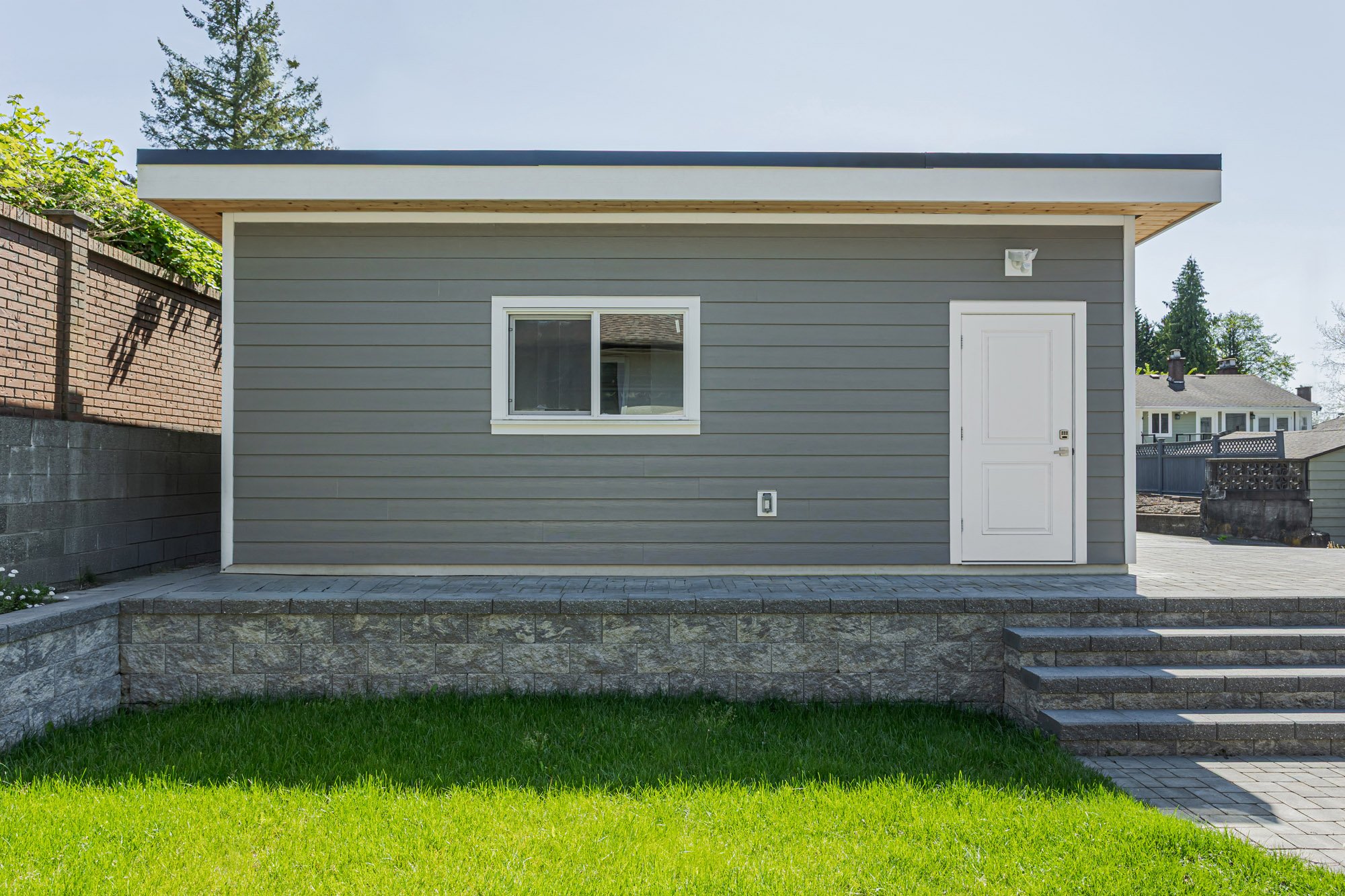 Back exterior view of a grey garage featuring a small window and a white door, sitting on a block retaining wall over grass.