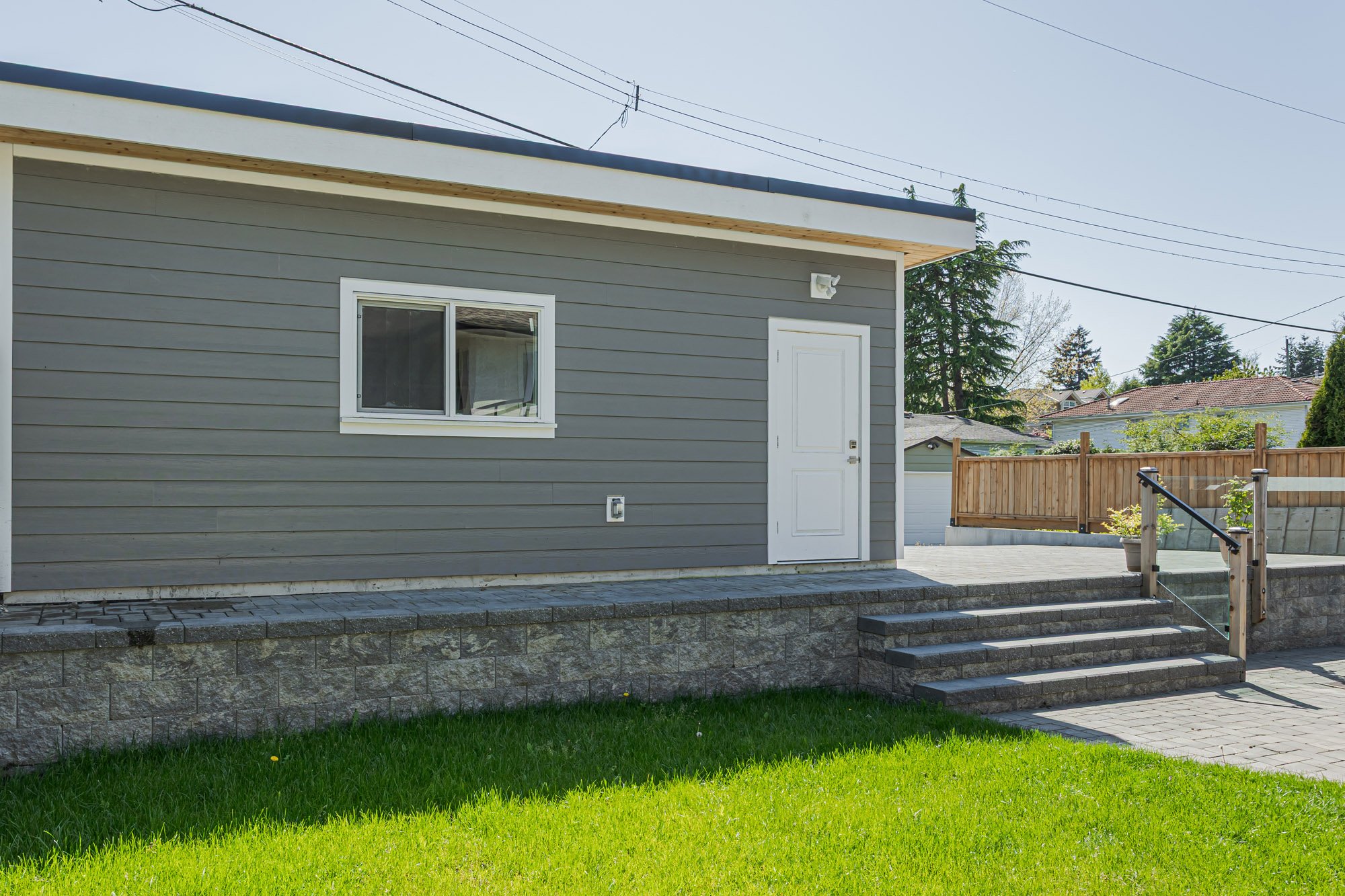 Angled rear view of a grey detached garage with a white side door and window, set beside a paved stone patio and a green lawn.