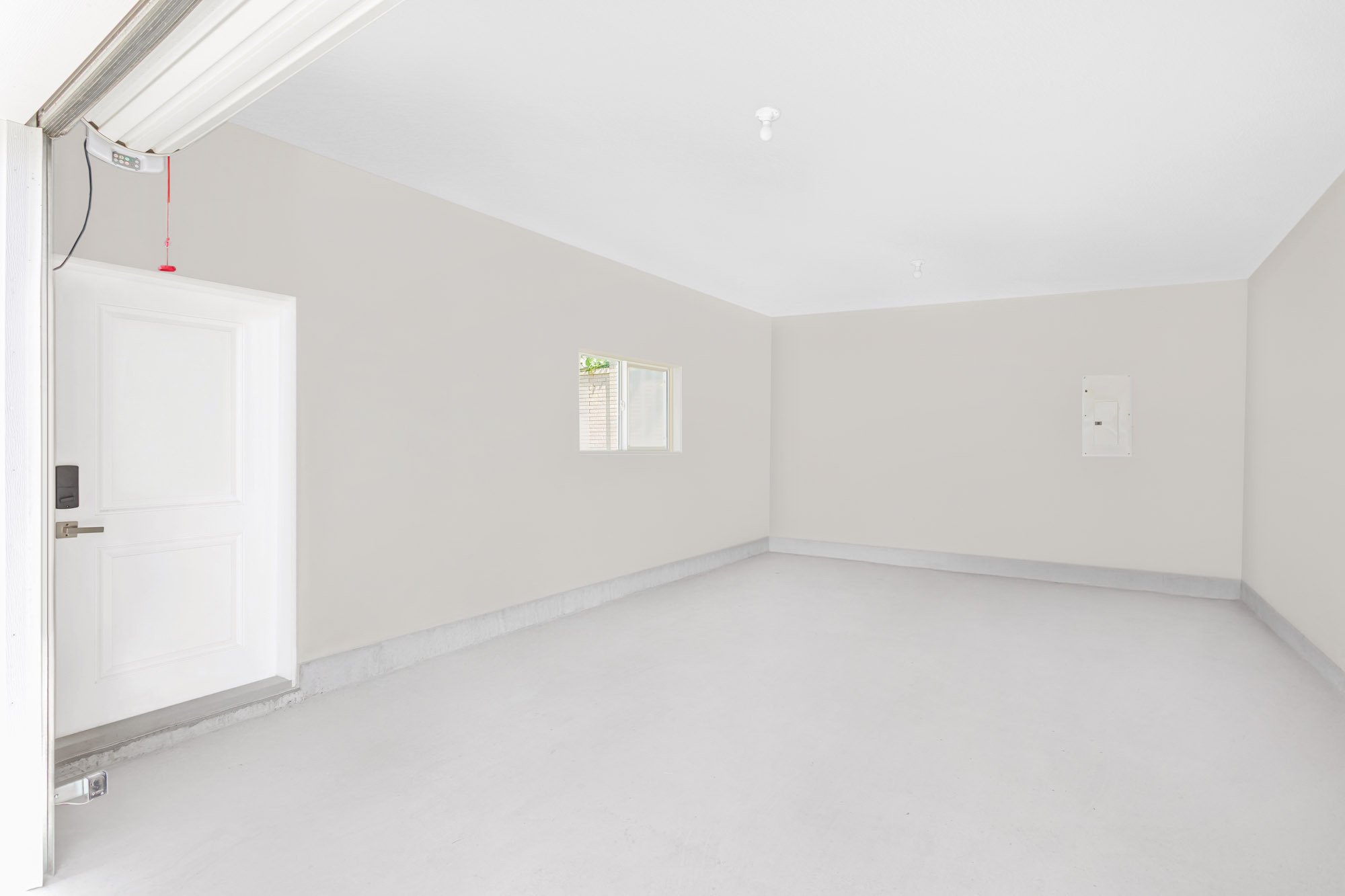 Bright, empty garage interior featuring a smooth light grey floor, beige walls, a white side entry door, and a small window.