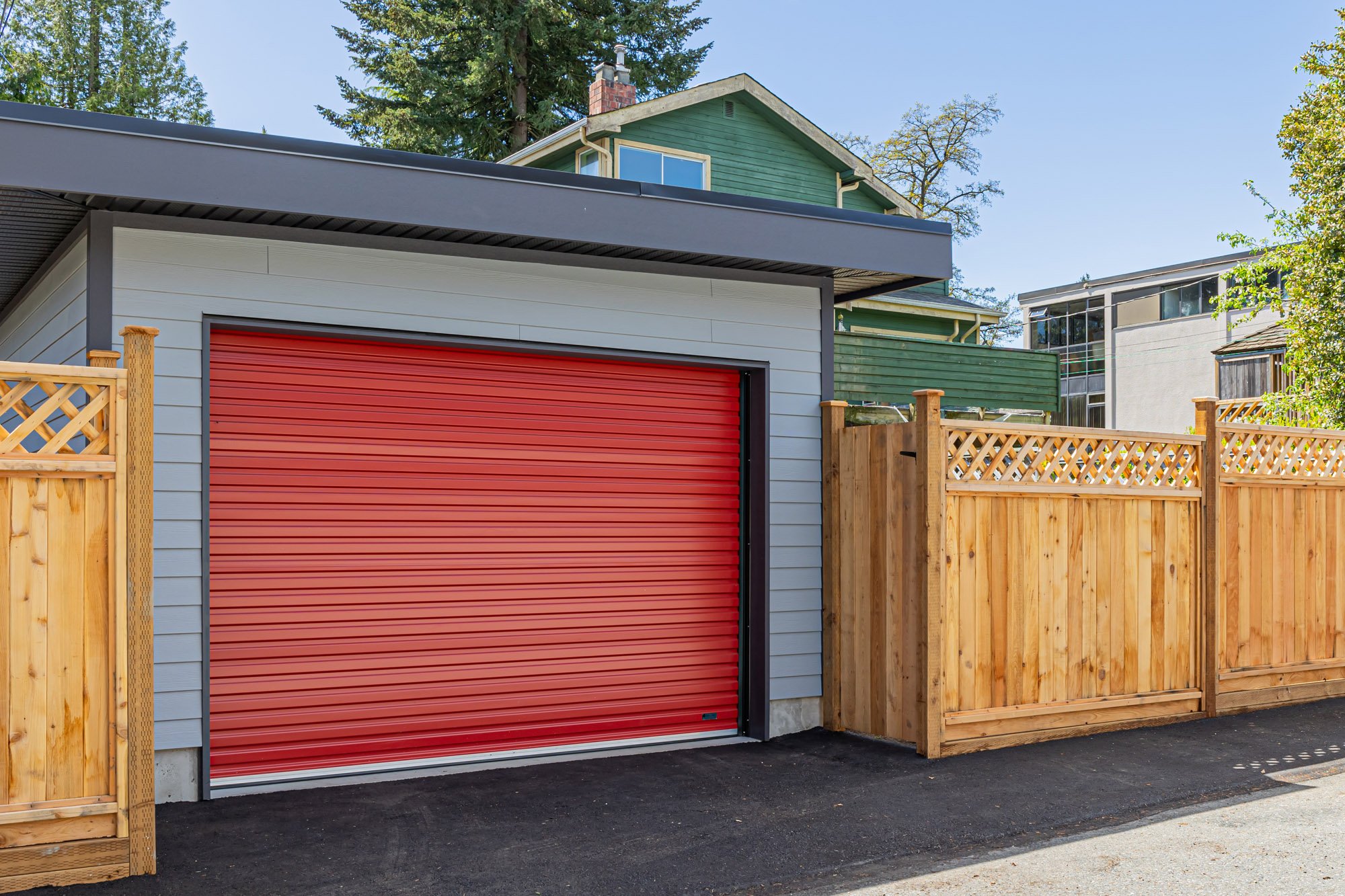 Angled front view of a modern grey garage with a bright red closed roll-up door and dark flat roof beside a wooden fence.