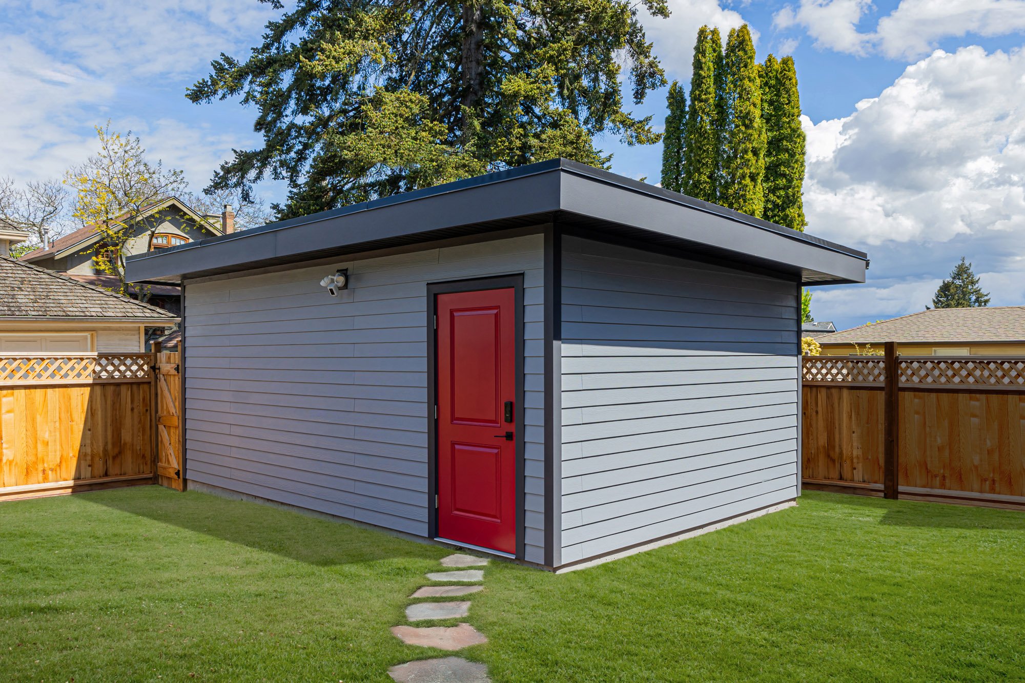 A modern detached grey garage with a flat roof and a vibrant red side door, surrounded by a green lawn and stepping stones.