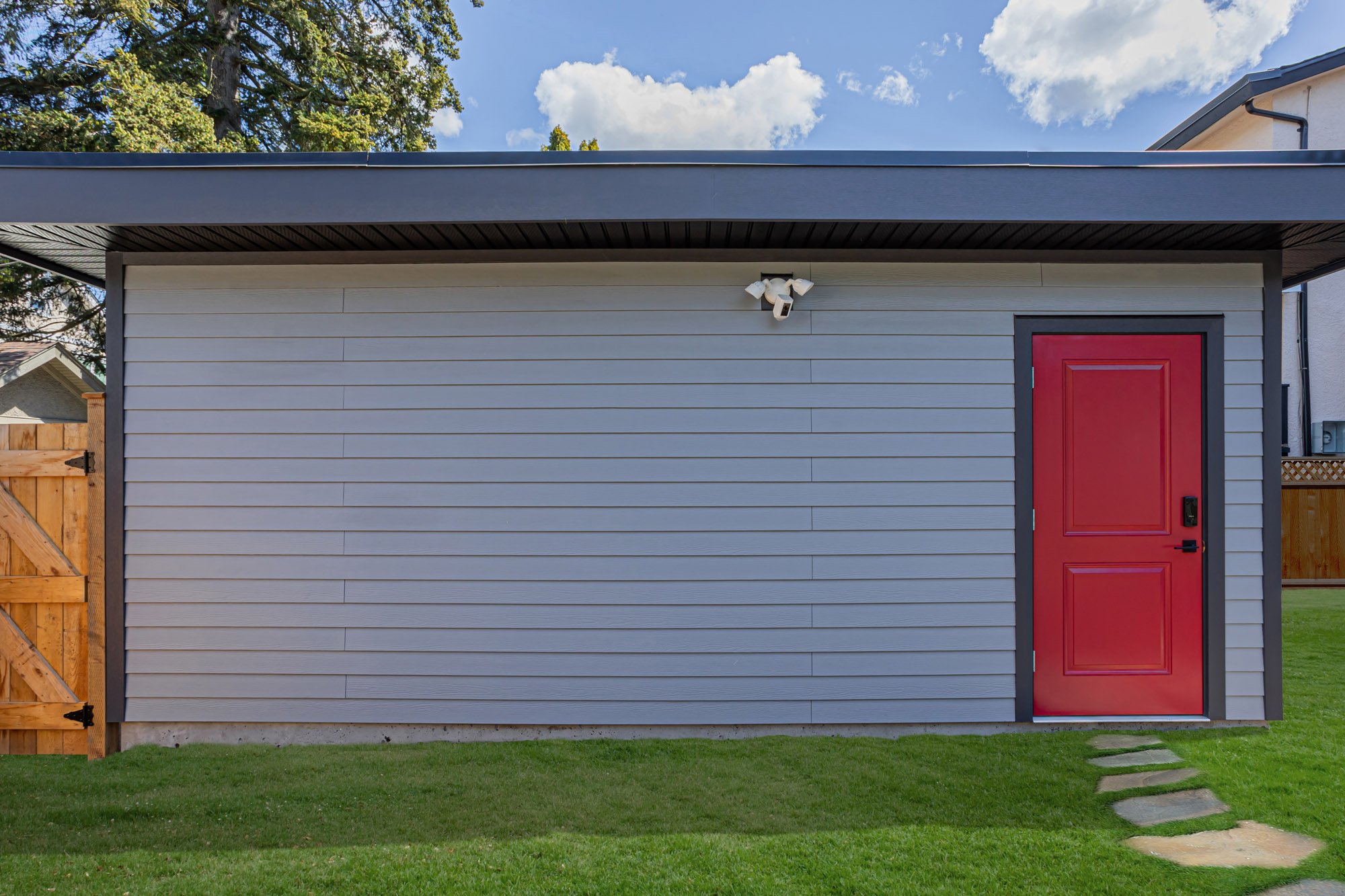 Straight-on exterior view of a modern grey garage featuring horizontal siding, a bright red side door, and a security light.