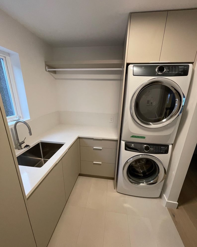 A bright laundry room featuring a white countertop with a stainless steel sink next to a stacked washer and dryer unit.