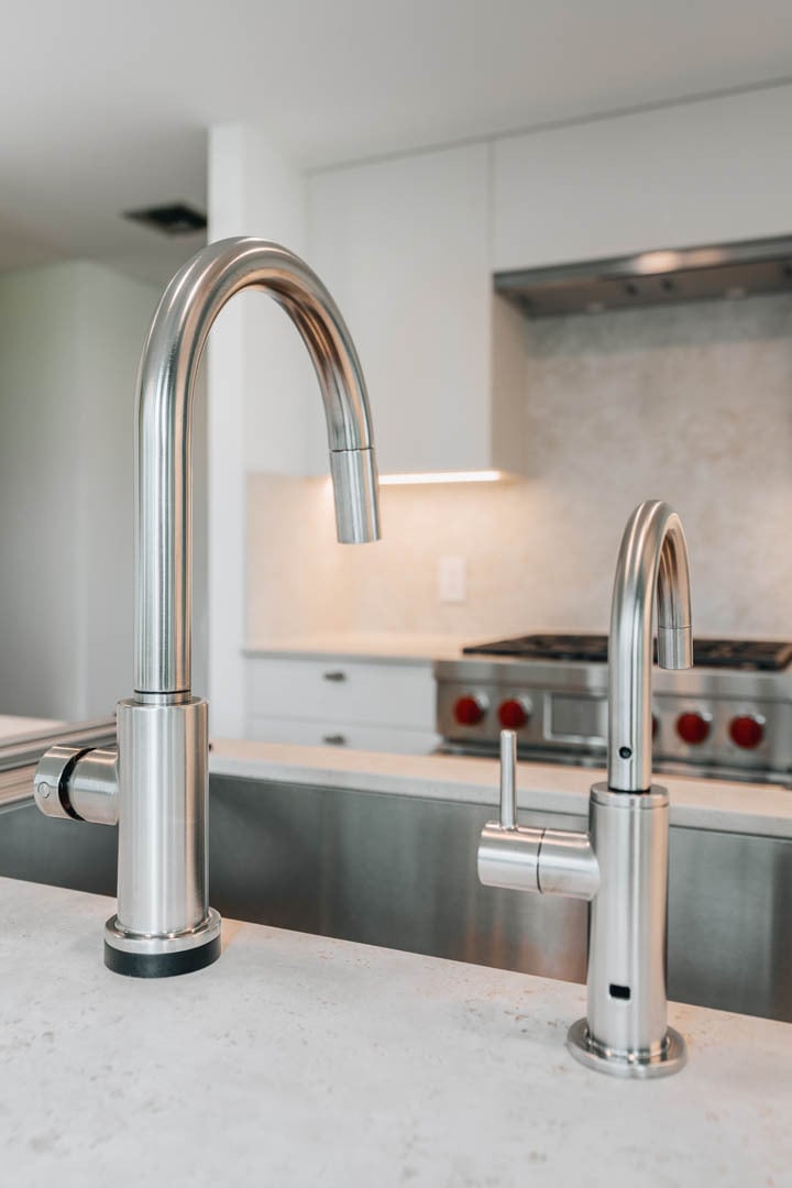 Close-up of two sleek stainless steel kitchen faucets, one large and one small, mounted on a bright white quartz countertop.