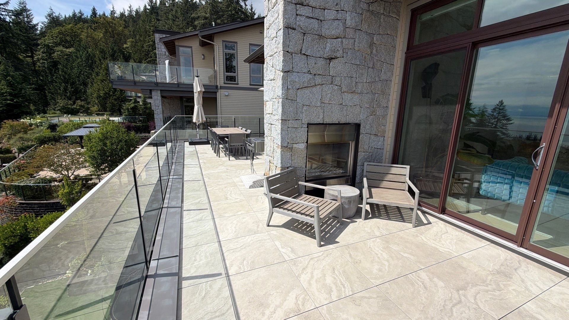 A spacious stone patio featuring glass railings, a built-in stone fireplace, two lounge chairs, and sliding glass doors.
