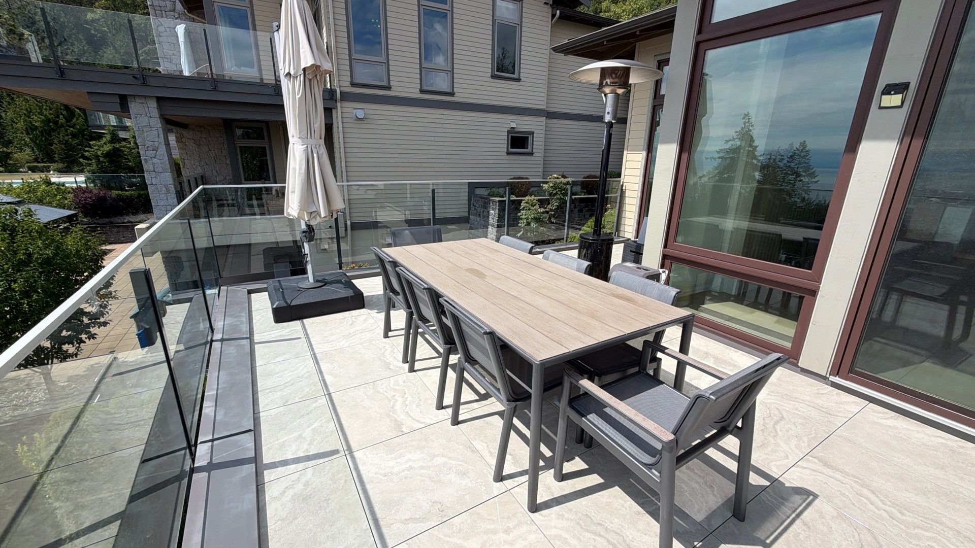 A sunny outdoor stone patio featuring a large dining table with grey chairs, a closed patio umbrella, and glass railings.