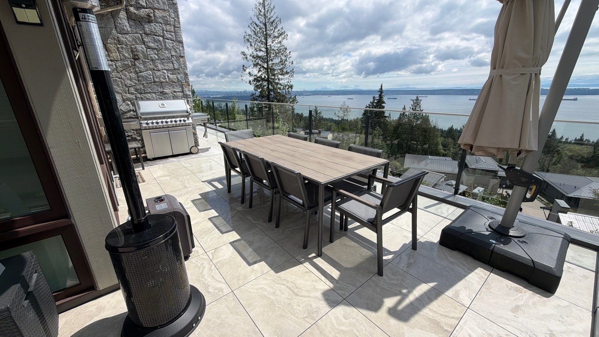 A spacious stone patio with an outdoor dining table, heater, and gas grill overlooking a stunning ocean view with ships.