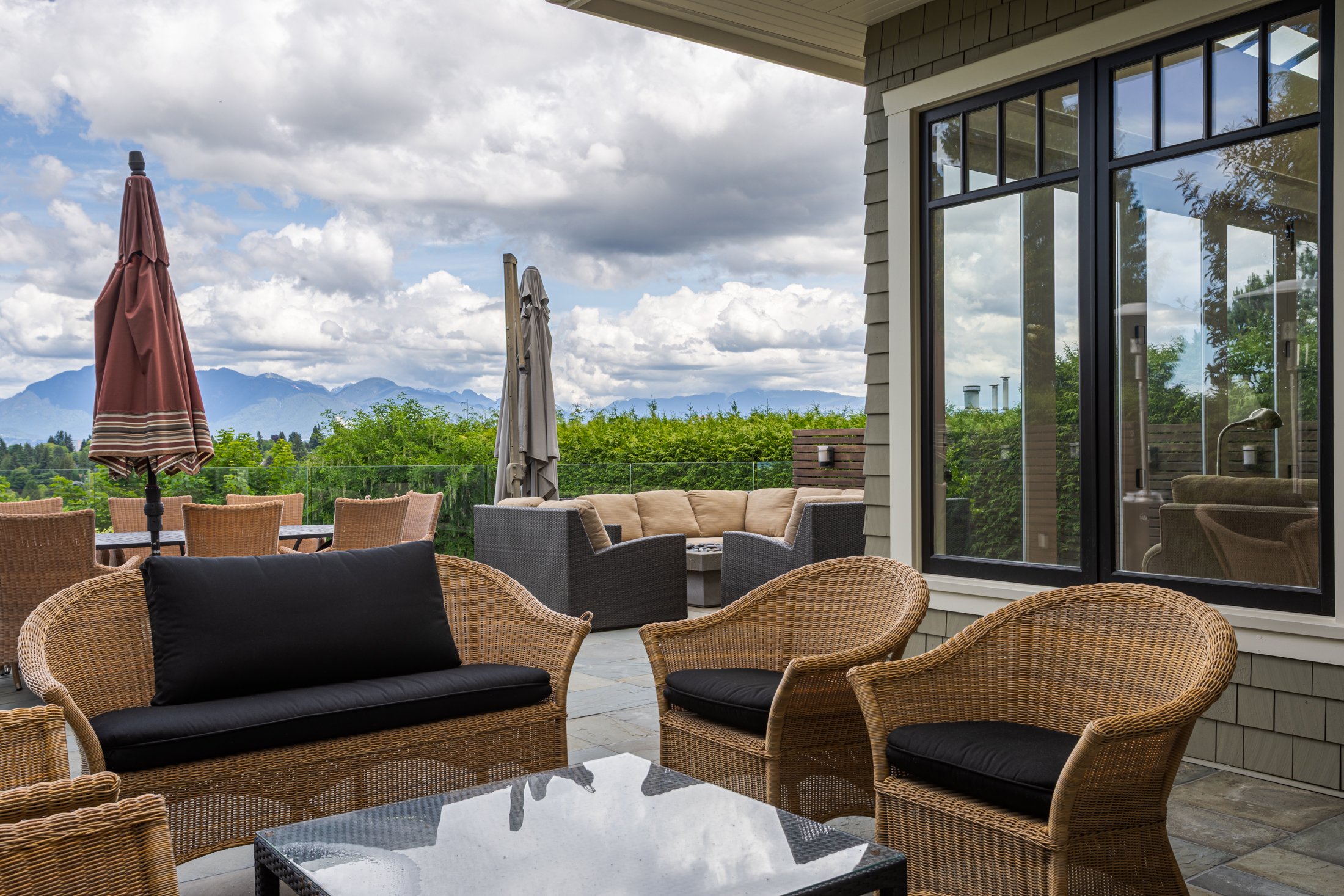 A covered outdoor patio featuring wicker seating with black cushions, large glass windows, and a distant mountain landscape.