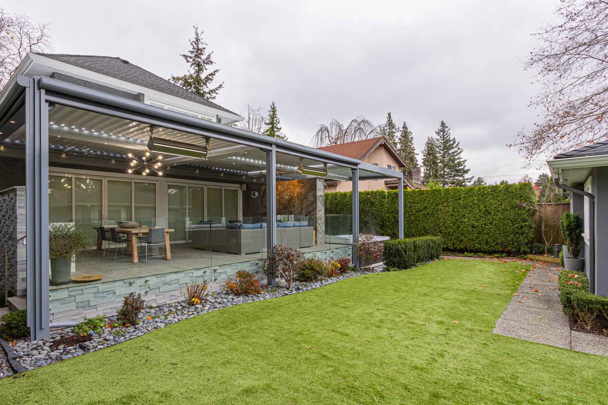 Wide view of a lush green backyard featuring a large covered patio with glass railings, outdoor seating, and tall hedges.