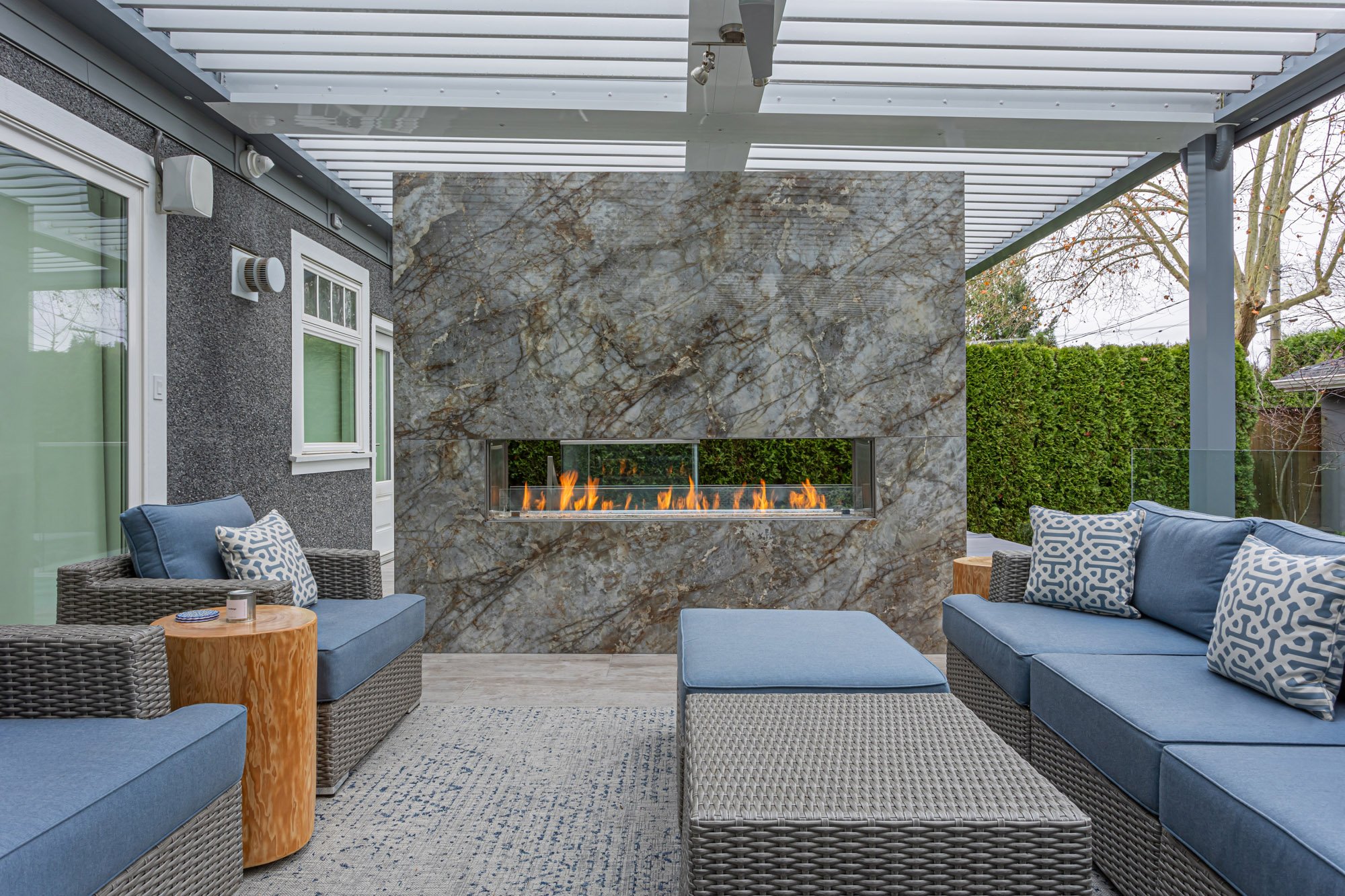 A covered outdoor living area featuring comfortable blue wicker seating arranged around a modern stone-clad gas fireplace.