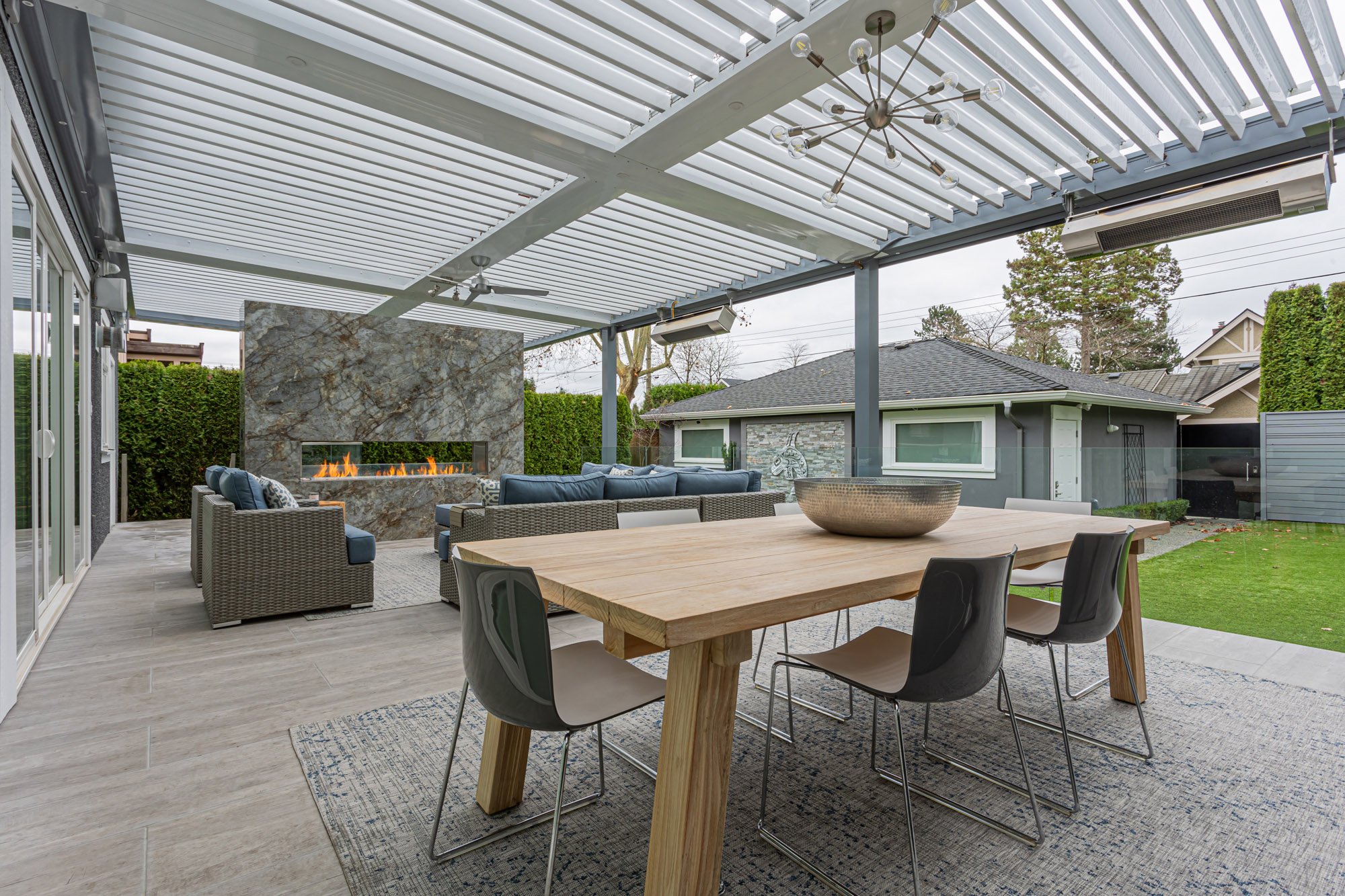 A covered outdoor living space featuring a wood dining table, a blue sectional sofa, and a striking stone gas fireplace.