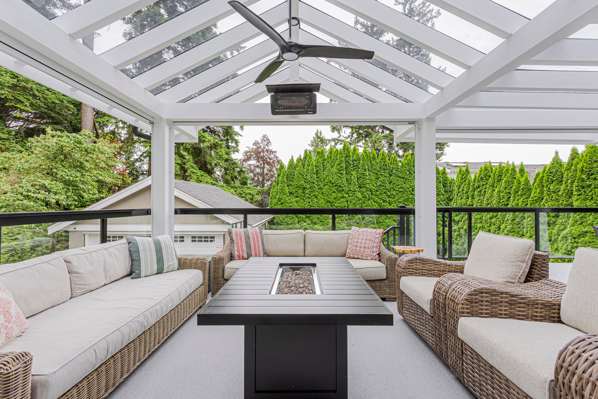 An inviting covered deck lounge area under a glass roof, featuring wicker seating, a black fire pit, and overhead heaters.